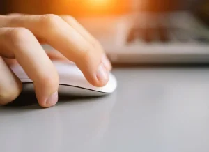 A close-up of a hand using a white computer mouse on a gray surface, with a laptop keyboard visible in the background. The image is warmly lit from the top left corner.