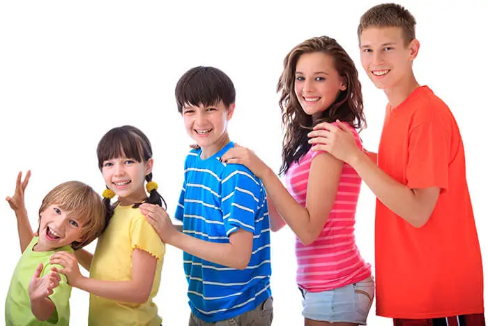 Five smiling children in colorful shirts stand in a line against a white background. They are leaning slightly forward, with each person resting their hands on the shoulders of the one in front.